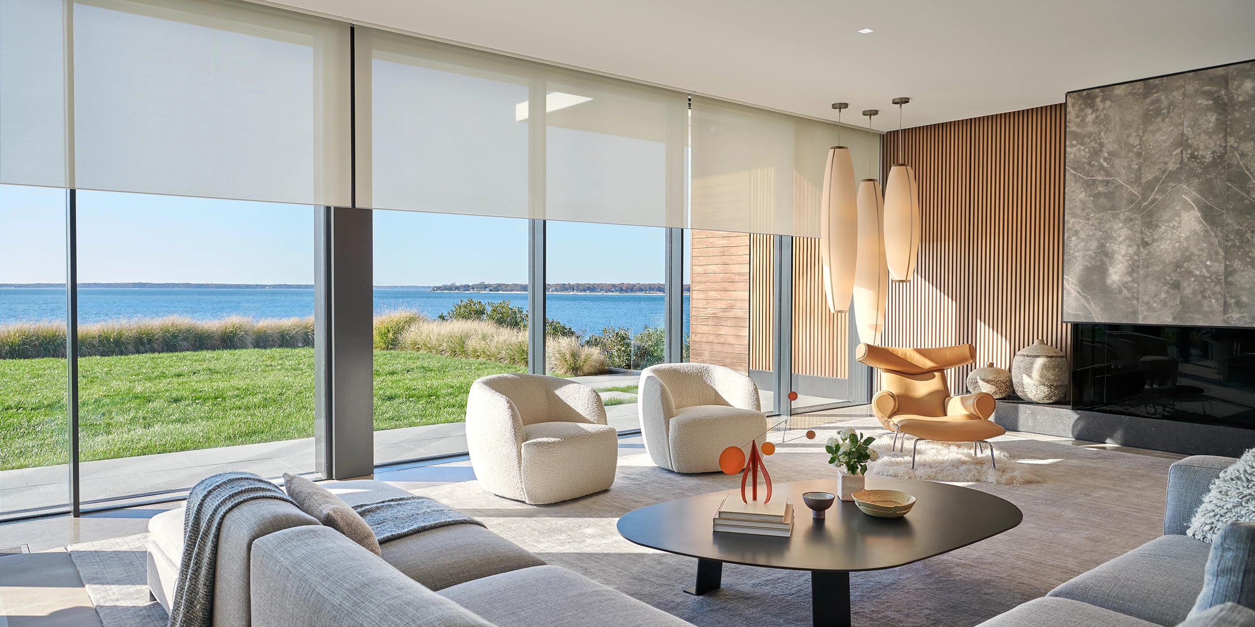 A contemporary living space with floor-to-ceiling windows, motorized shades, and elegant furniture, overlooking a waterfront landscape.
