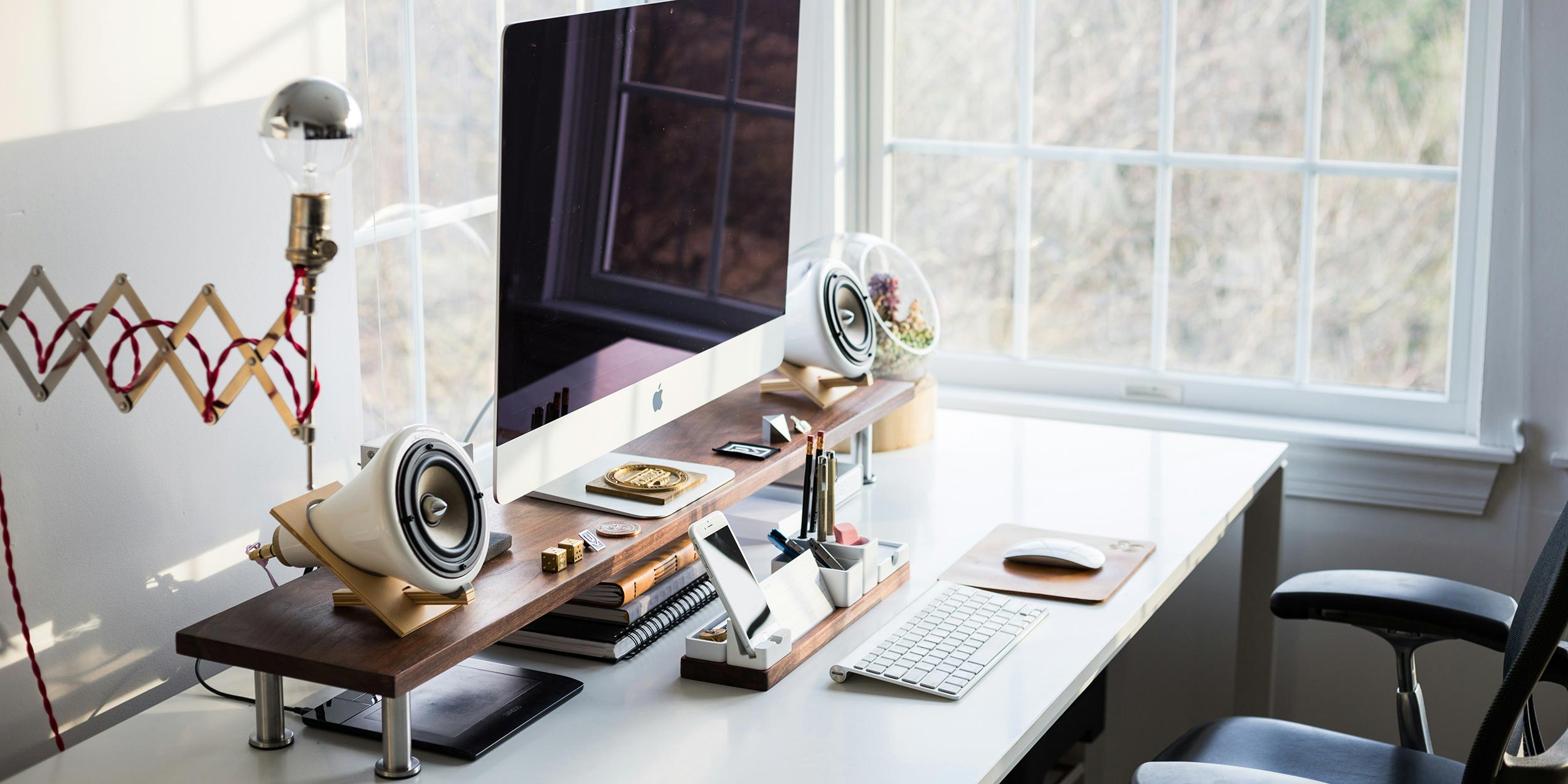 A stylish home office setup with an Apple computer, modern speakers, and neatly organized desk accessories in a bright, naturally lit room.